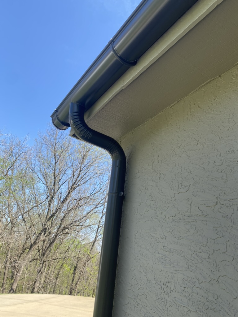 Black gutter and downspout installed on building with white stucco siding-leafless tree in distance against blue sky