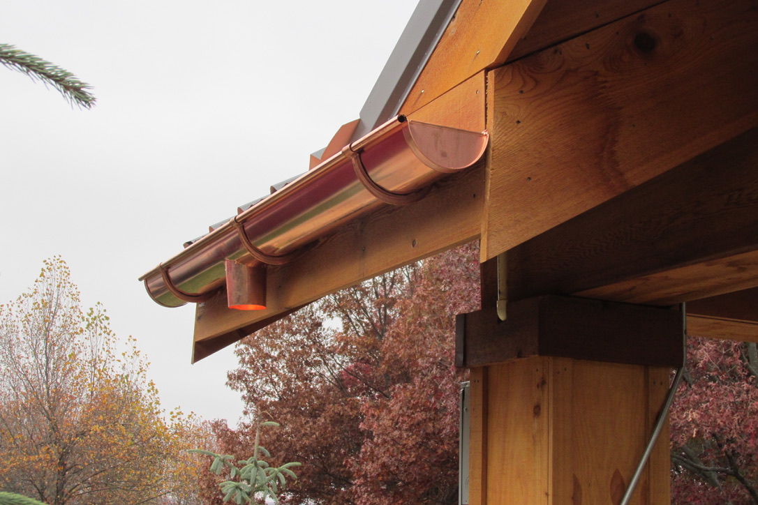 A close-up view of a copper gutter system installed on the edge of a wooden pole barn roof. The gutter has a curved profile and is securely fastened to the wooden eaves. The rich, natural wood of the barn contrasts with the warm tones of the copper. The background features autumn-colored trees under an overcast sky.