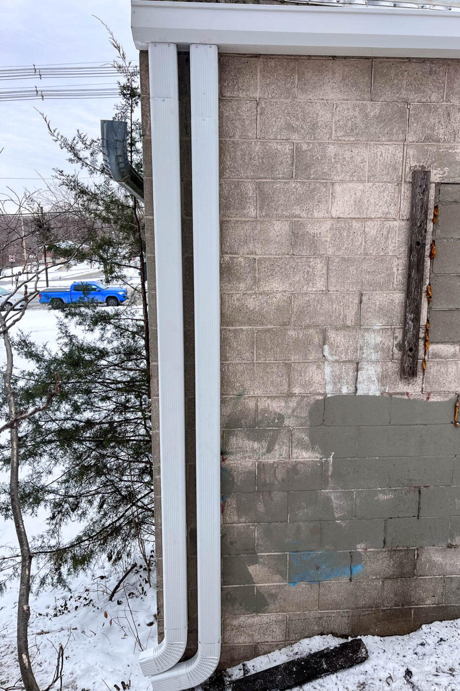 Overhead view of a 7-inch white commercial gutter system with a 4x5 downspout installed along a concrete block wall, with snow-covered ground and a blue truck visible in the background.