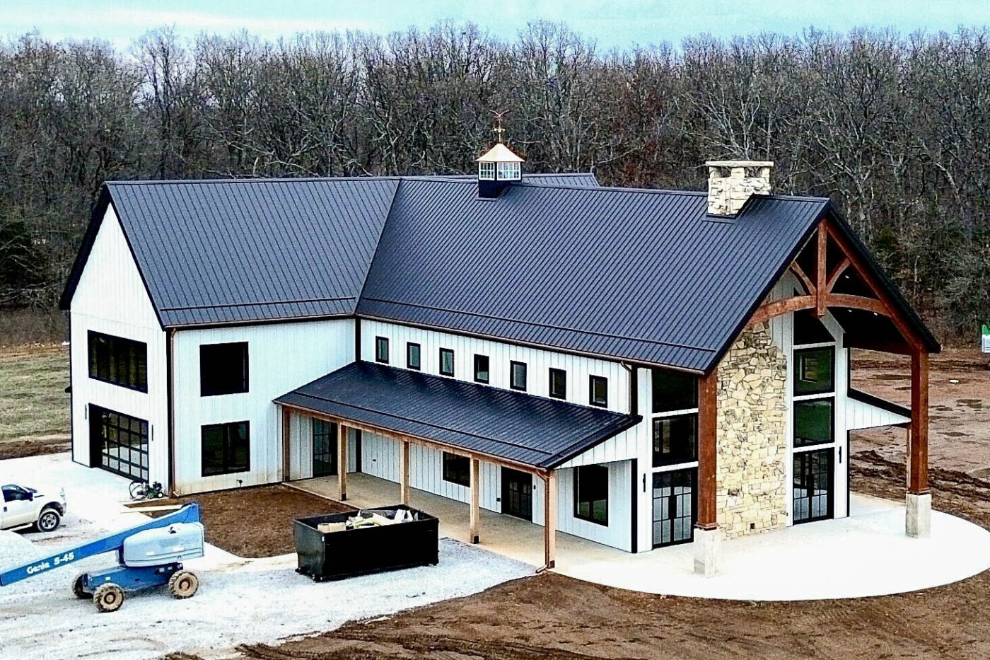 Wide view of a modern home with dark roofing, copper half-round gutters, and round copper downspouts, set in a wooded rural landscape during construction.