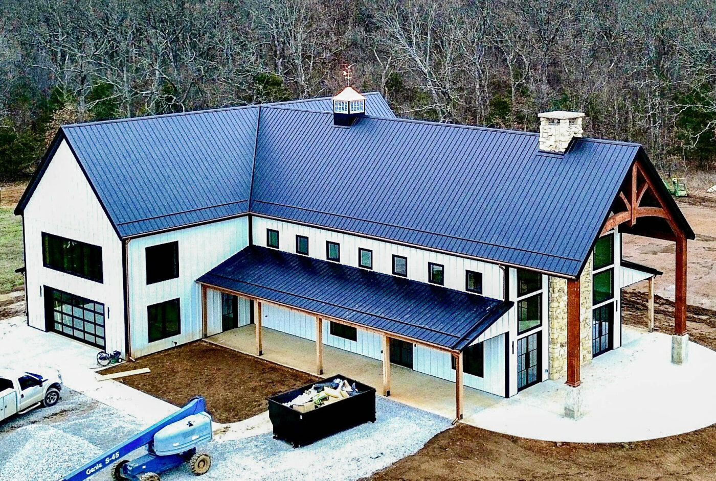 Aerial view of a large modern home with black metal roofing and half-round copper gutters installed along the roofline, surrounded by a wooded backdrop and construction equipment.