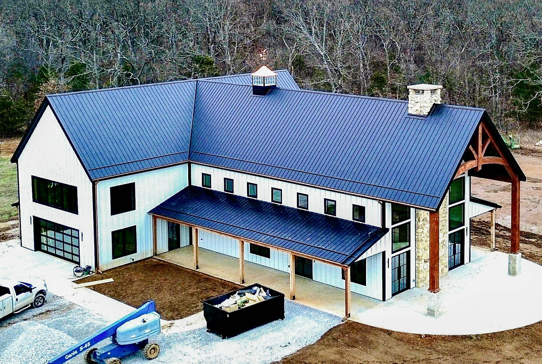 Aerial view of a large modern home with black metal roofing and half-round copper gutters installed along the roofline, surrounded by a wooded backdrop and construction equipment.
