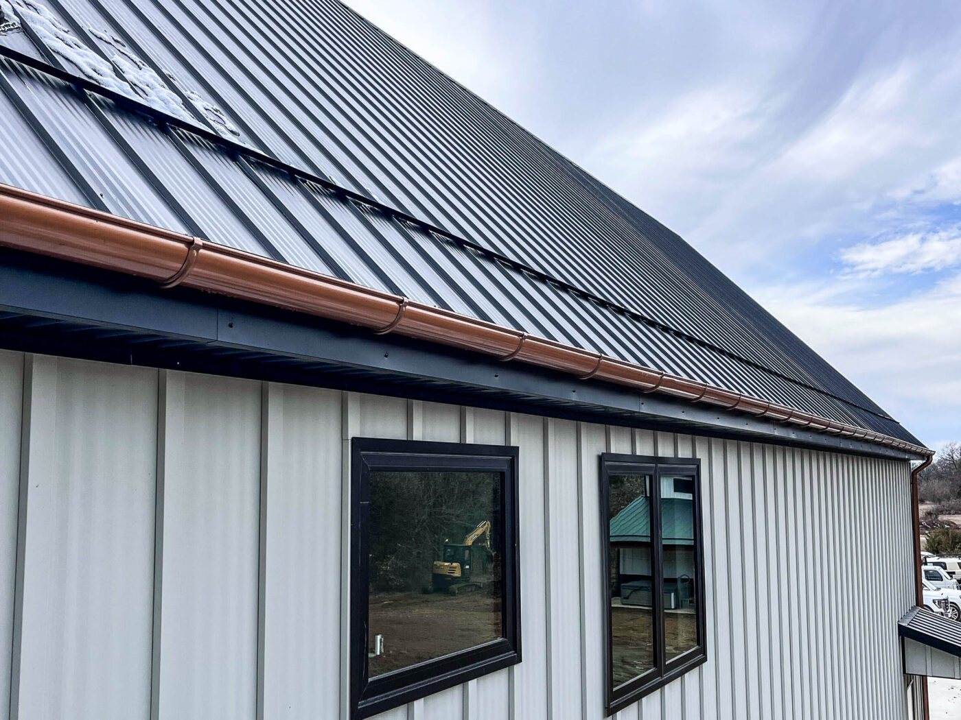Close-up view of a standing seam metal roof with 6-inch half-round copper gutters and a round copper downspout mounted on a light-colored modern exterior wall.