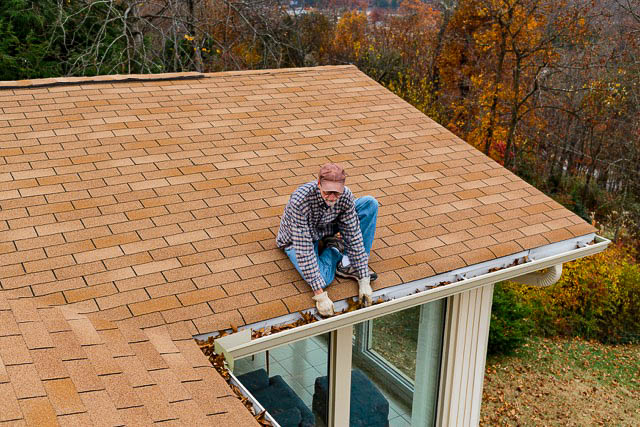 An elderly man sitting on a roof, manually cleaning out a heavily clogged home gutter.