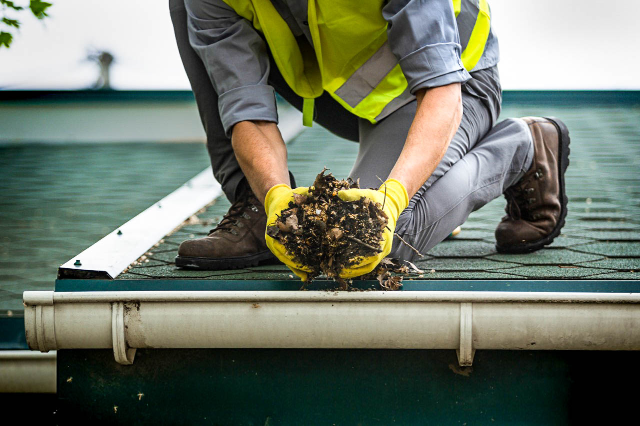 A worker wearing gloves and a high-visibility vest removing wet leaves and debris from a residential gutter.