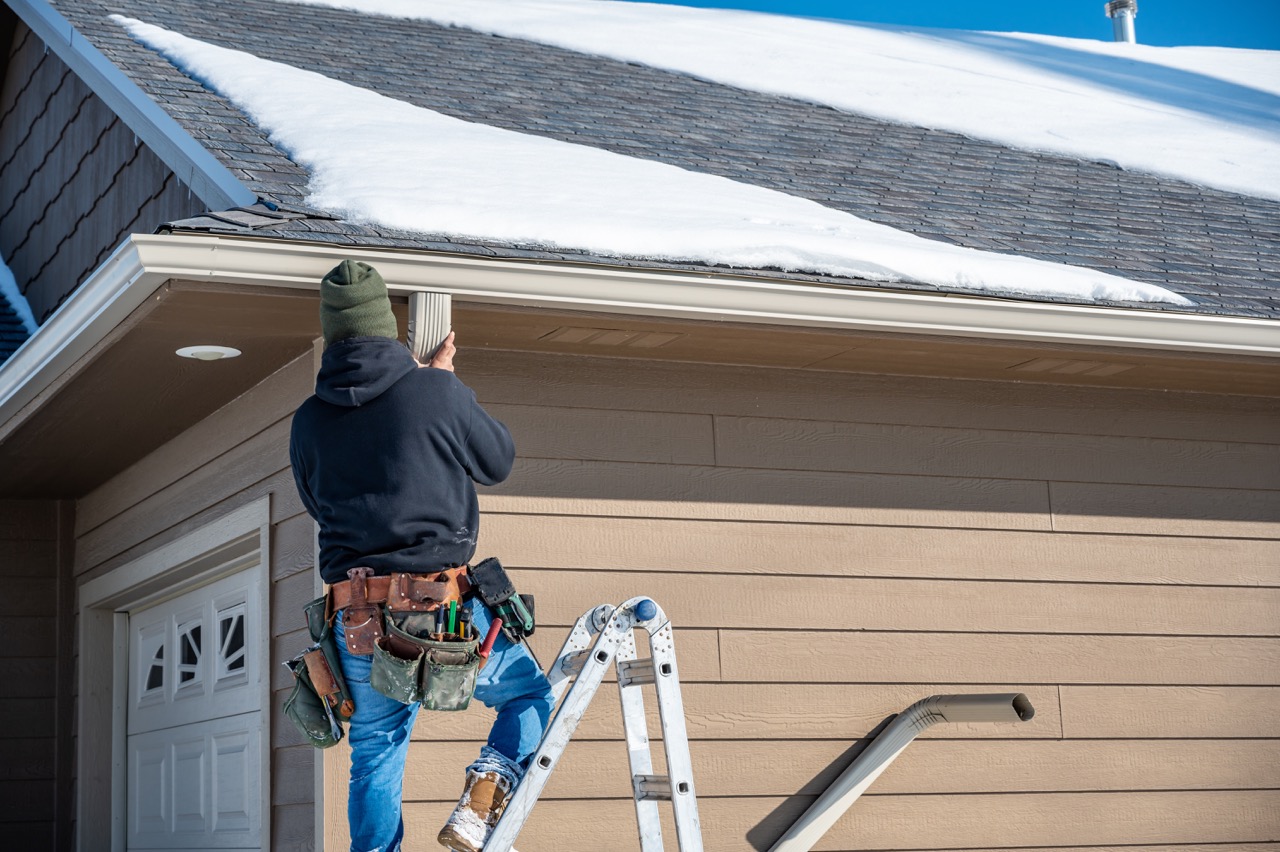 contractor installing gutters on a residential building in the winter with snow on the roof 