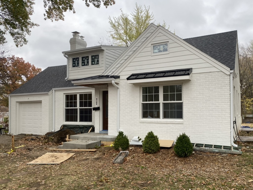 White gutters and downspouts installed on white house of brick and dark gray shingled roof-fallen brown leaves on ground