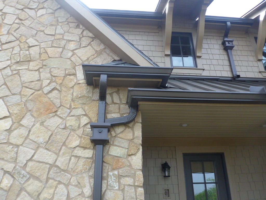 Black gutters and downspout of two-story beige stone and tile house