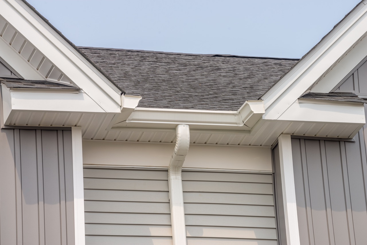 White gutter with downpipe on the dark gray roof of a gray house-Gutter Installation in Buckner MO